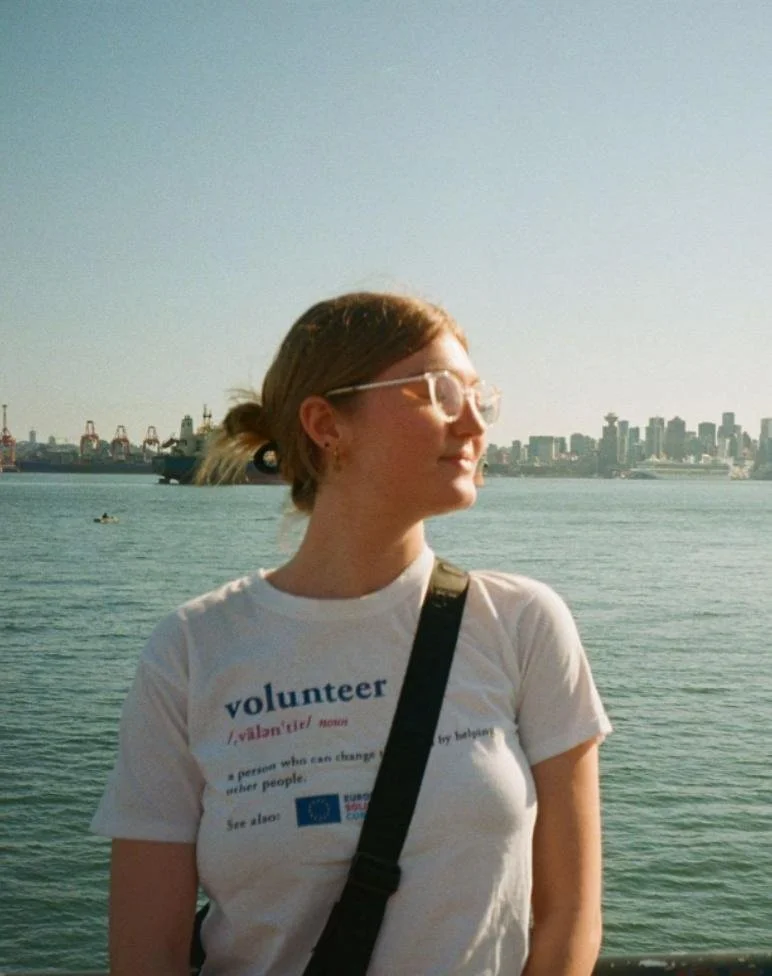 A woman standing by the water with a city skyline in the background, wearing glasses, a white T-shirt that says 'volunteer,' and a black strap across her shoulder.