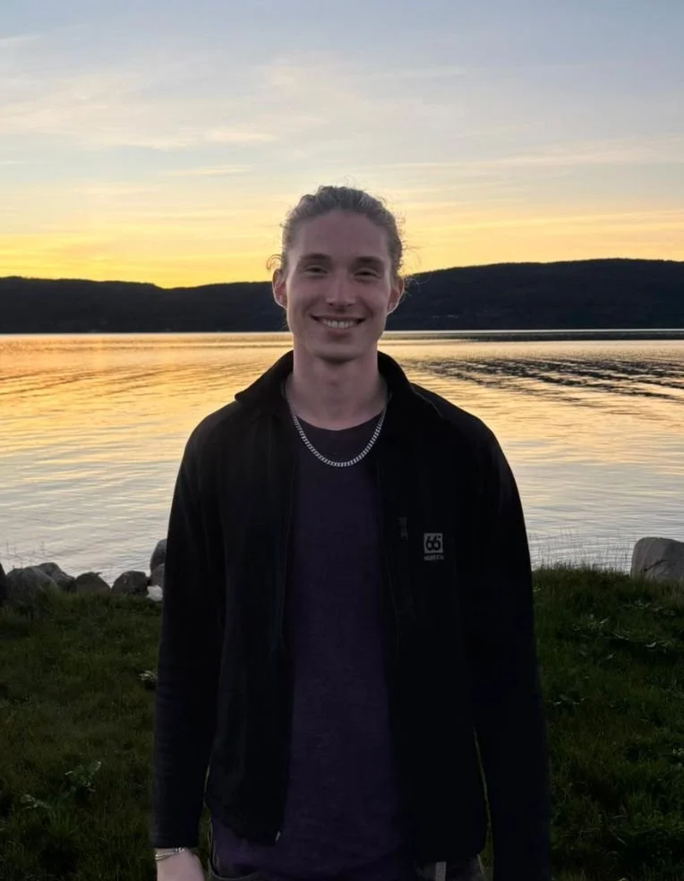 A young man standing by a lake during sunset, smiling at the camera, wearing a black jacket and a silver chain, with hills and a colorful sky reflected in the water.