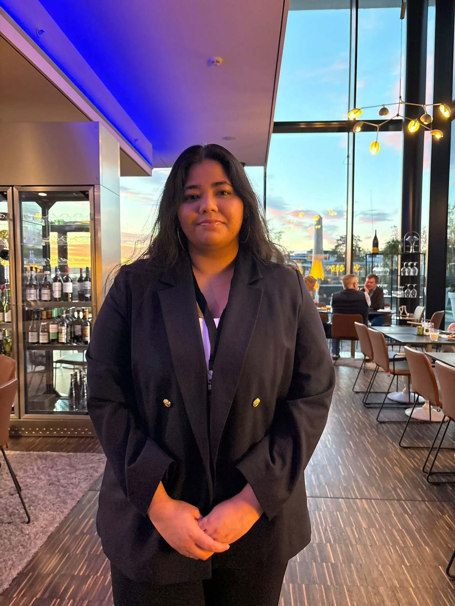 A woman standing inside a modern restaurant with large floor-to-ceiling windows during sunset.