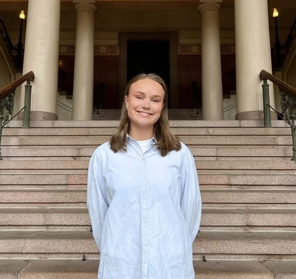 A young woman with light brown hair, wearing a white coat, standing on the steps of a building with large columns and ornate railings.