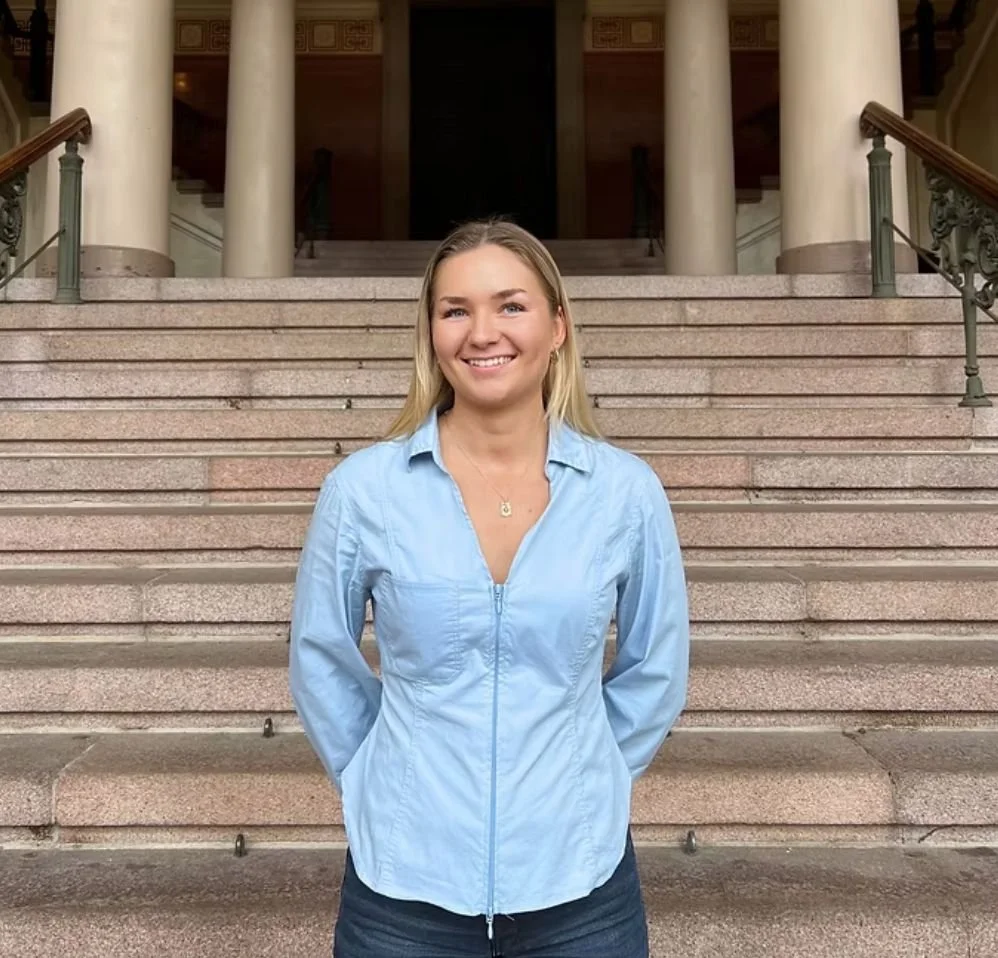 A smiling young woman standing on stone steps outside a building with tall columns.