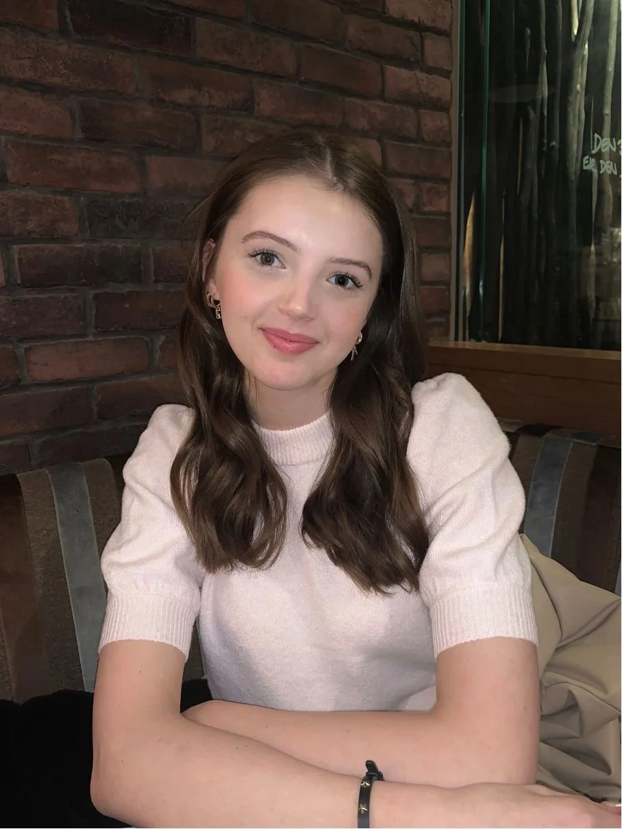 Young woman with wavy brown hair, wearing a light pink short-sleeve sweater, sitting at a table with a brick wall background.