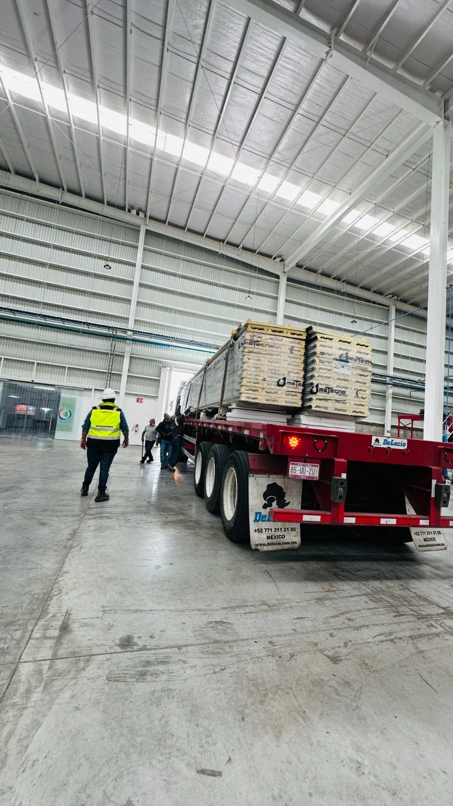 Camión en una nave industrial con varias personas trabajando, uno con chaleco de seguridad amarillo y cascos, cargando materiales de construcción.