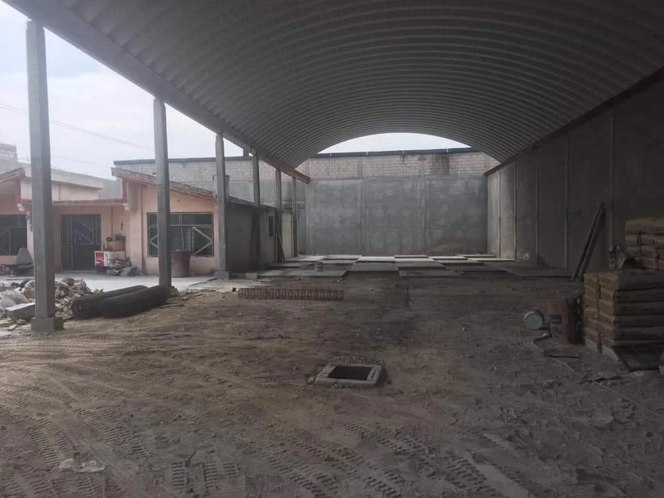 Estructura en construcción en un espacio cerrado con techo metálico curvado y paredes de concreto, con materiales de construcción y tierra en el suelo.