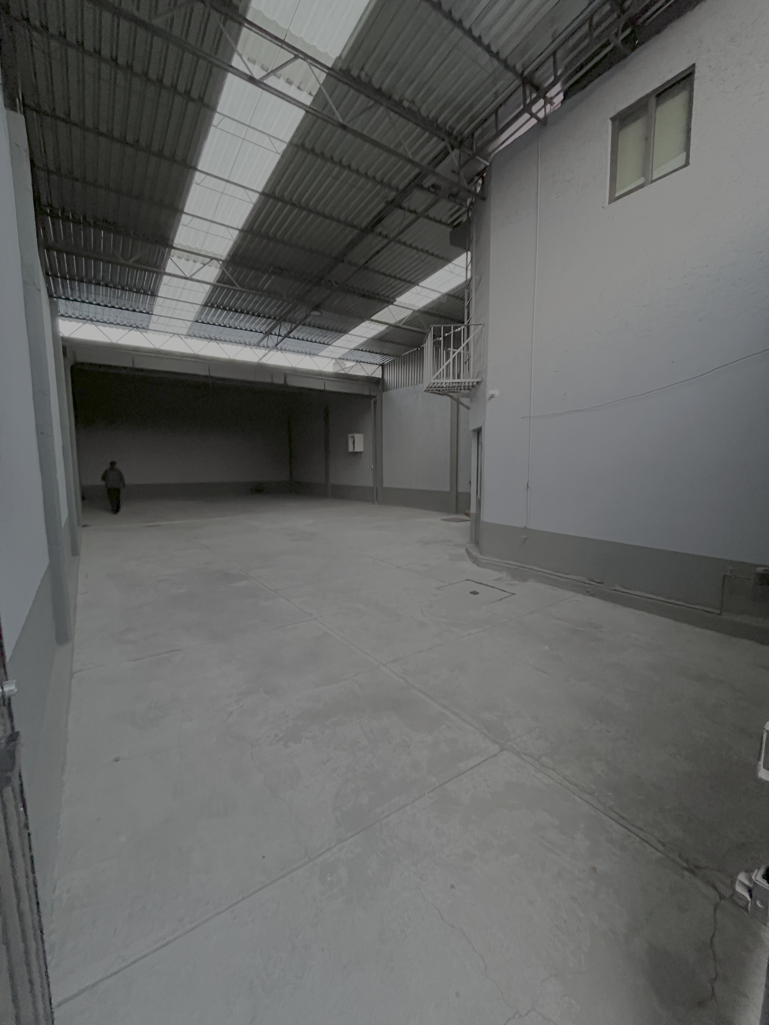 Espacio interior de un almacén o salón grande con techos altos de lámina metálica y paredes blancas. Tiene una superficie de concreto y poca iluminación natural.