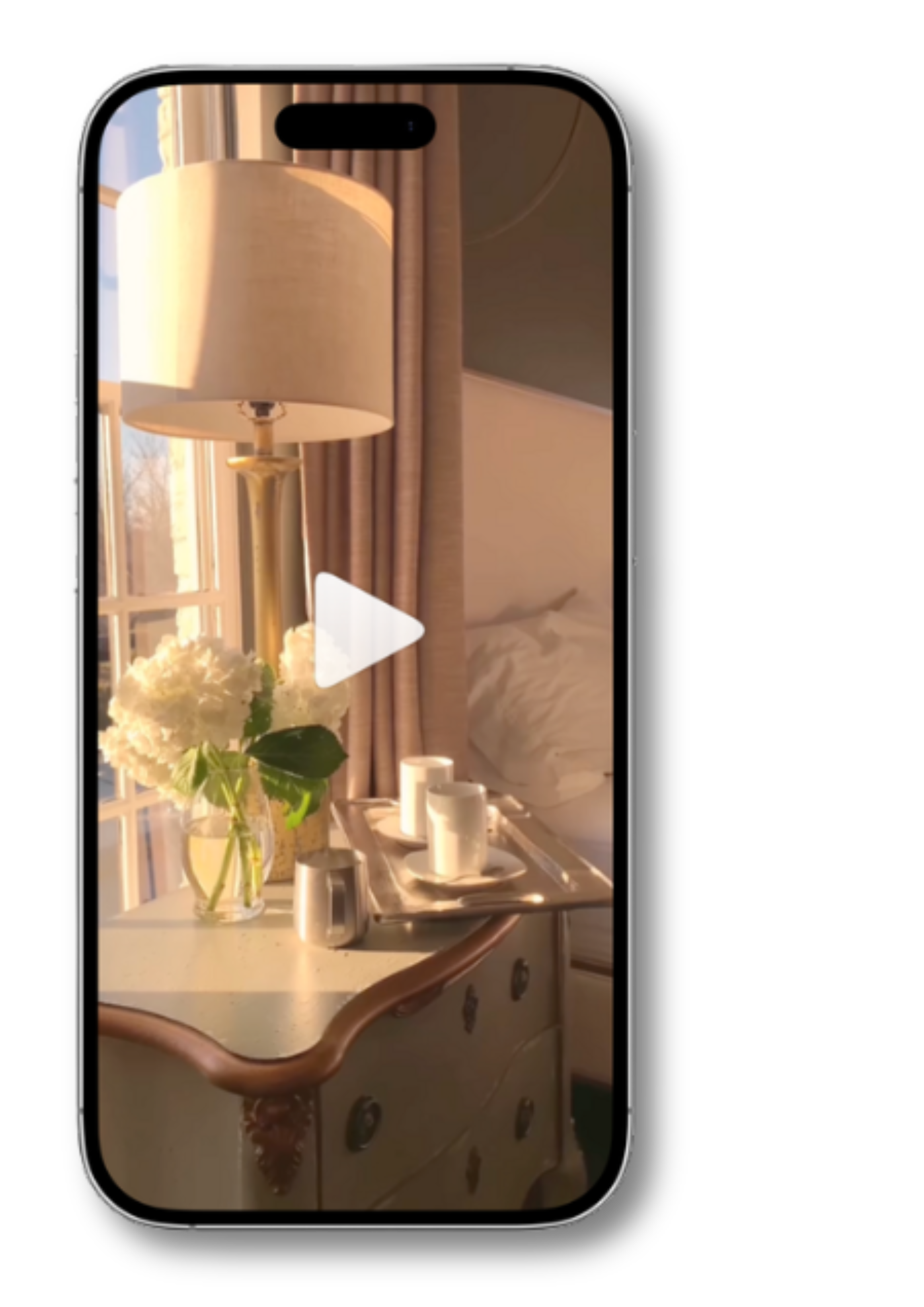 Smartphone screen displaying a video of a bedside table with a lamp, white flowers in a glass vase, and a tray with cups, near a bed with white bedding and beige curtains.