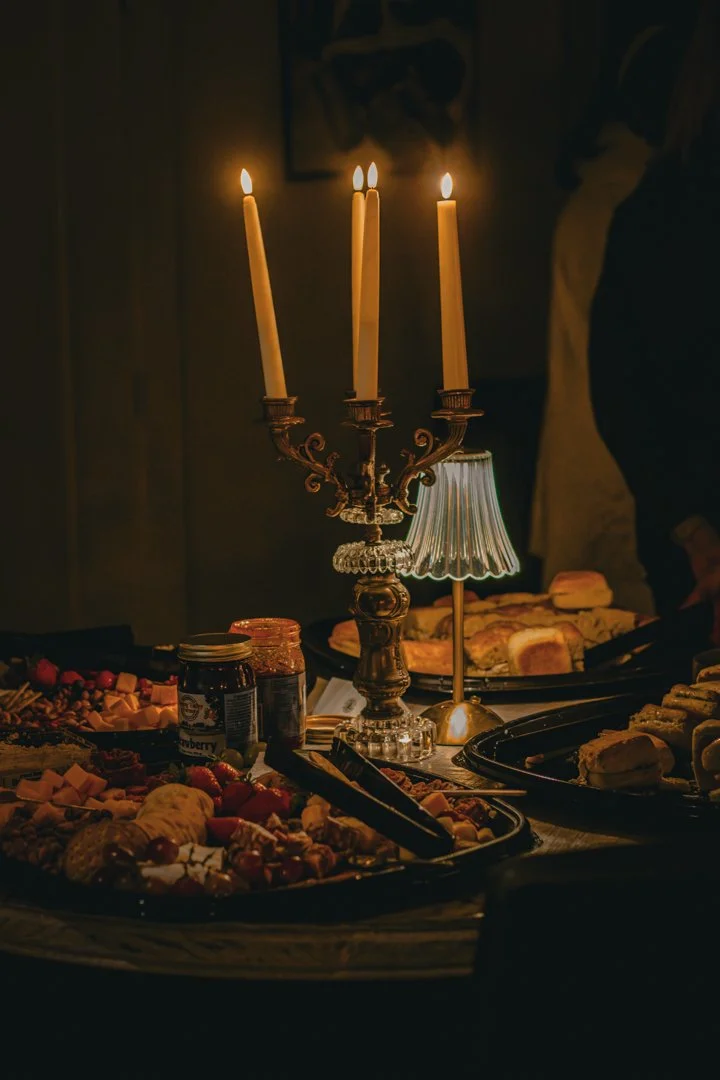 A dimly lit table with a candelabra holding three lit candles, a small table lamp, and various desserts including cookies, fruit, and jam.