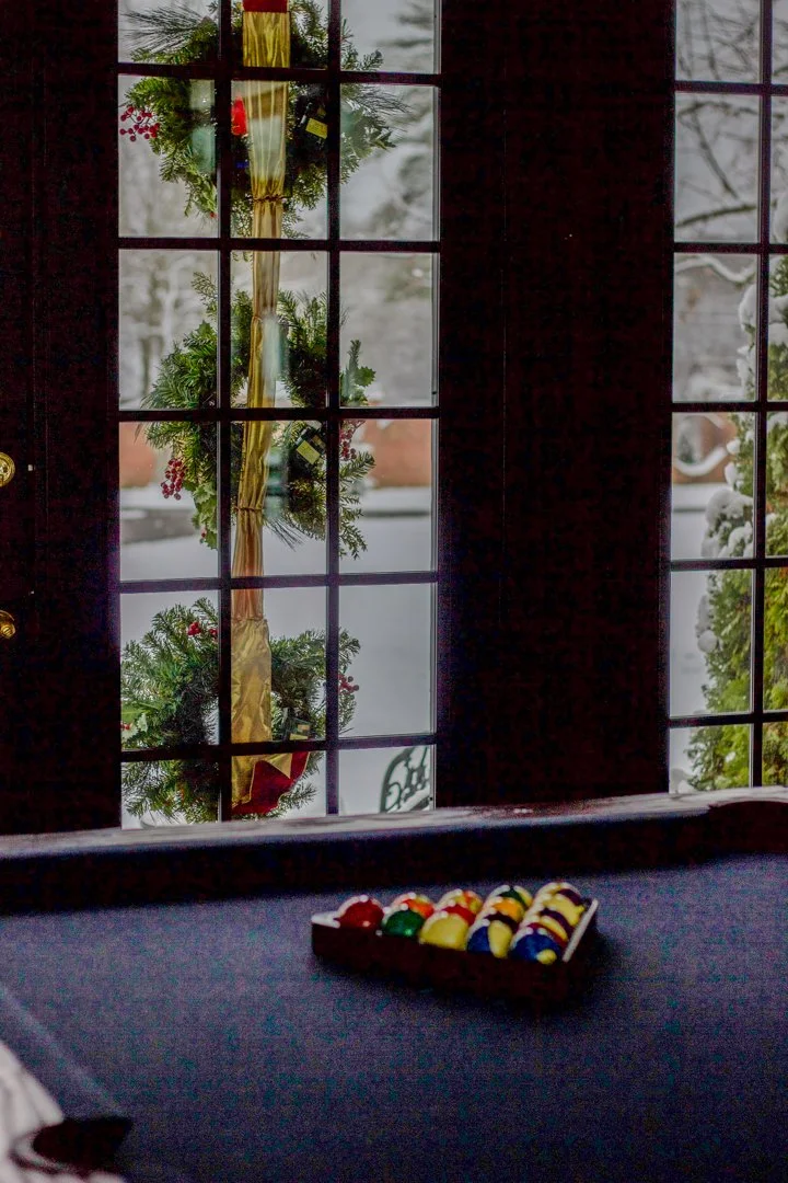 A view from inside a house looking out through glass-paneled doors decorated for Christmas with a wreath. Snow covers the ground outside. On a table inside, there is a tray of colorful golf balls.
