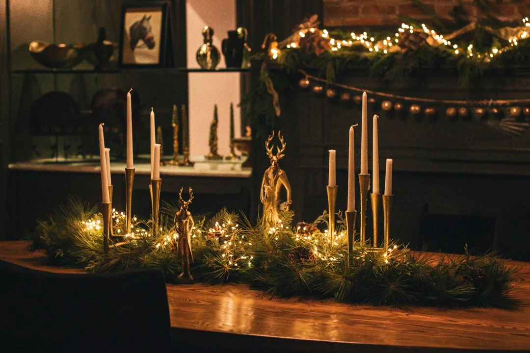 A holiday table centerpiece with candles, gold reindeer figurines, pine branches, and fairy lights, set in a dimly lit room with Christmas decorations.