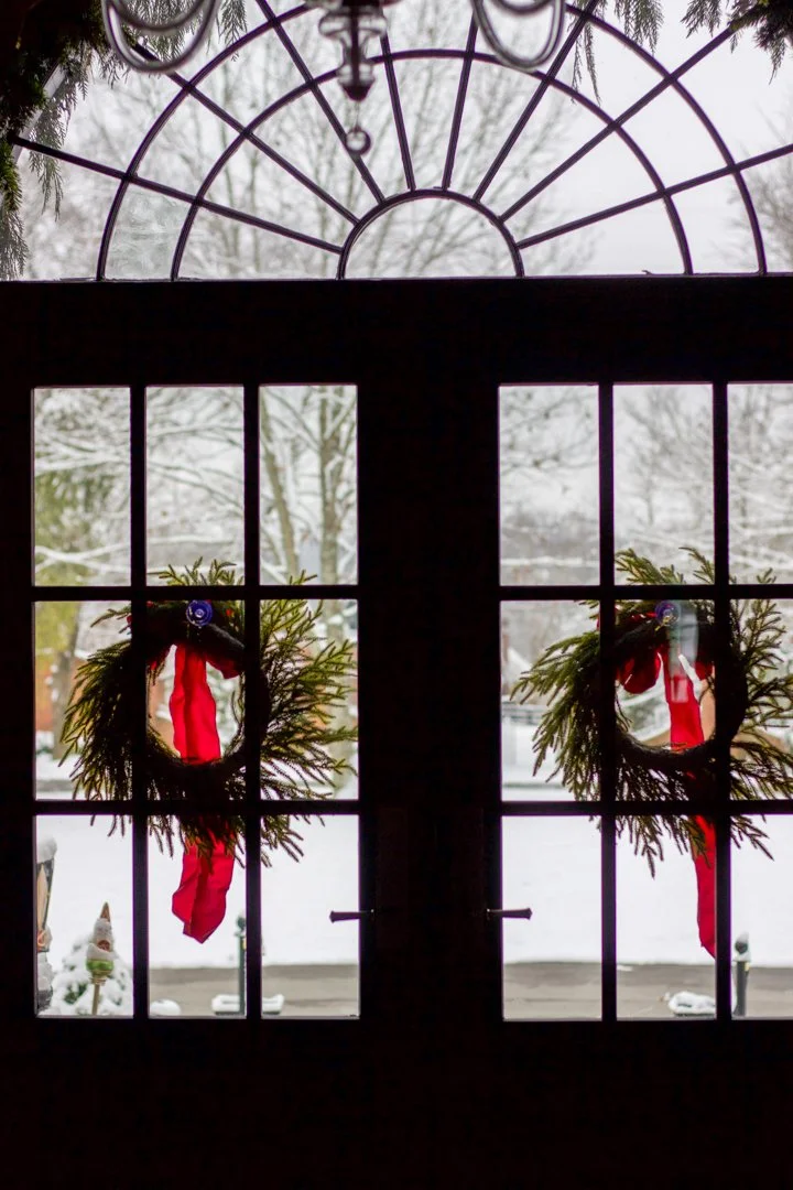 View through decorated window showing snow-covered landscape, wreaths with red ribbons on window, and small snowman outside.