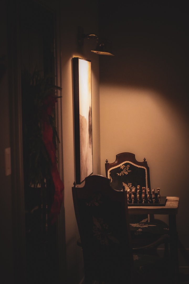 Dimly lit room with a wall-mounted lamp, a painting on the wall, and a vintage-style table with a chessboard and chess pieces, with an ornate chair in front.
