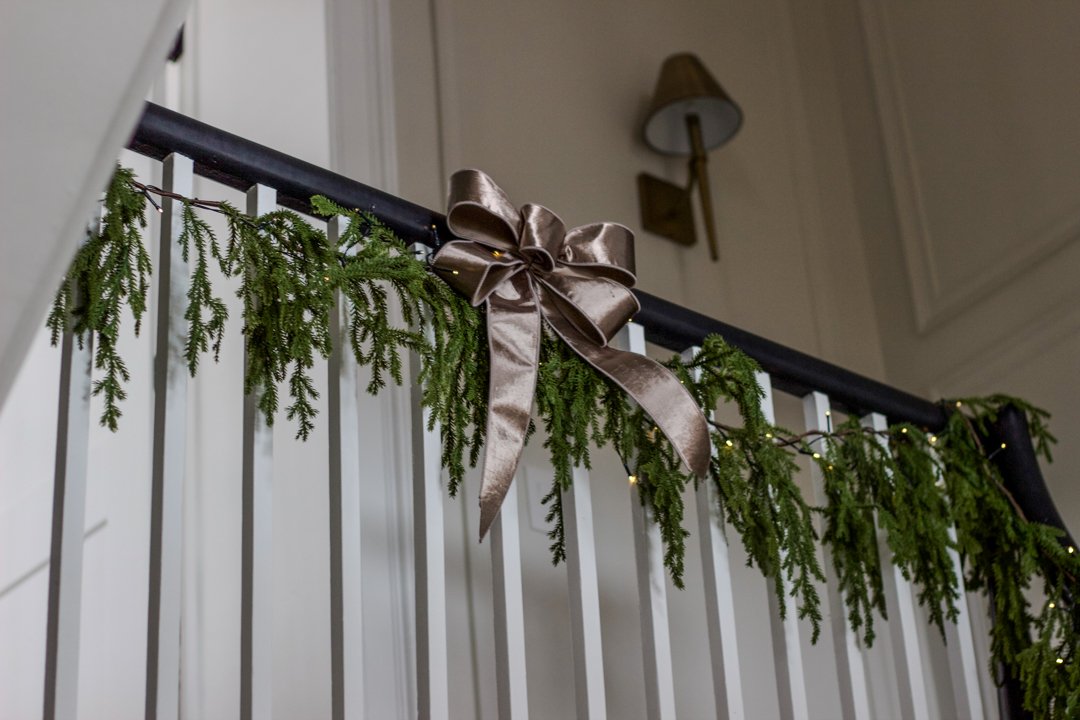 Decorated staircase with green garland and a large metallic ribbon bow, with a wall-mounted lamp in the background.