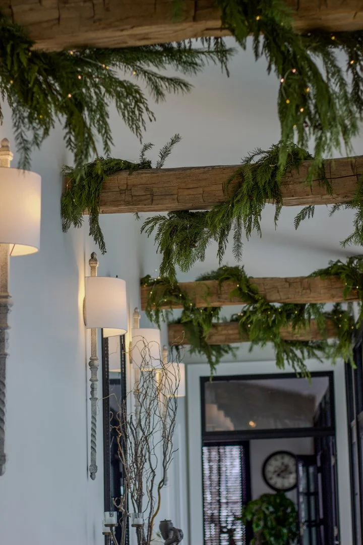 Indoor hallway decorated with greenery and wooden beams on the ceiling, illuminated by wall-mounted lamps, with a clock and window in the background.
