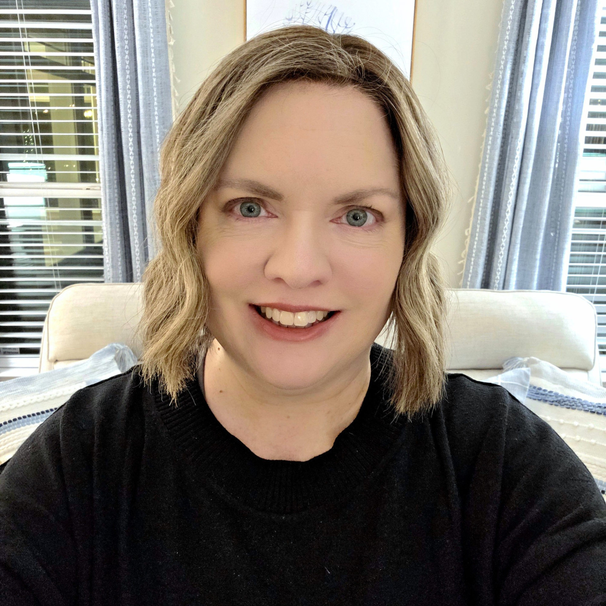 Close-up of a smiling woman with blonde wavy hair, wearing a black top, sitting in a room with large windows and blue curtains.