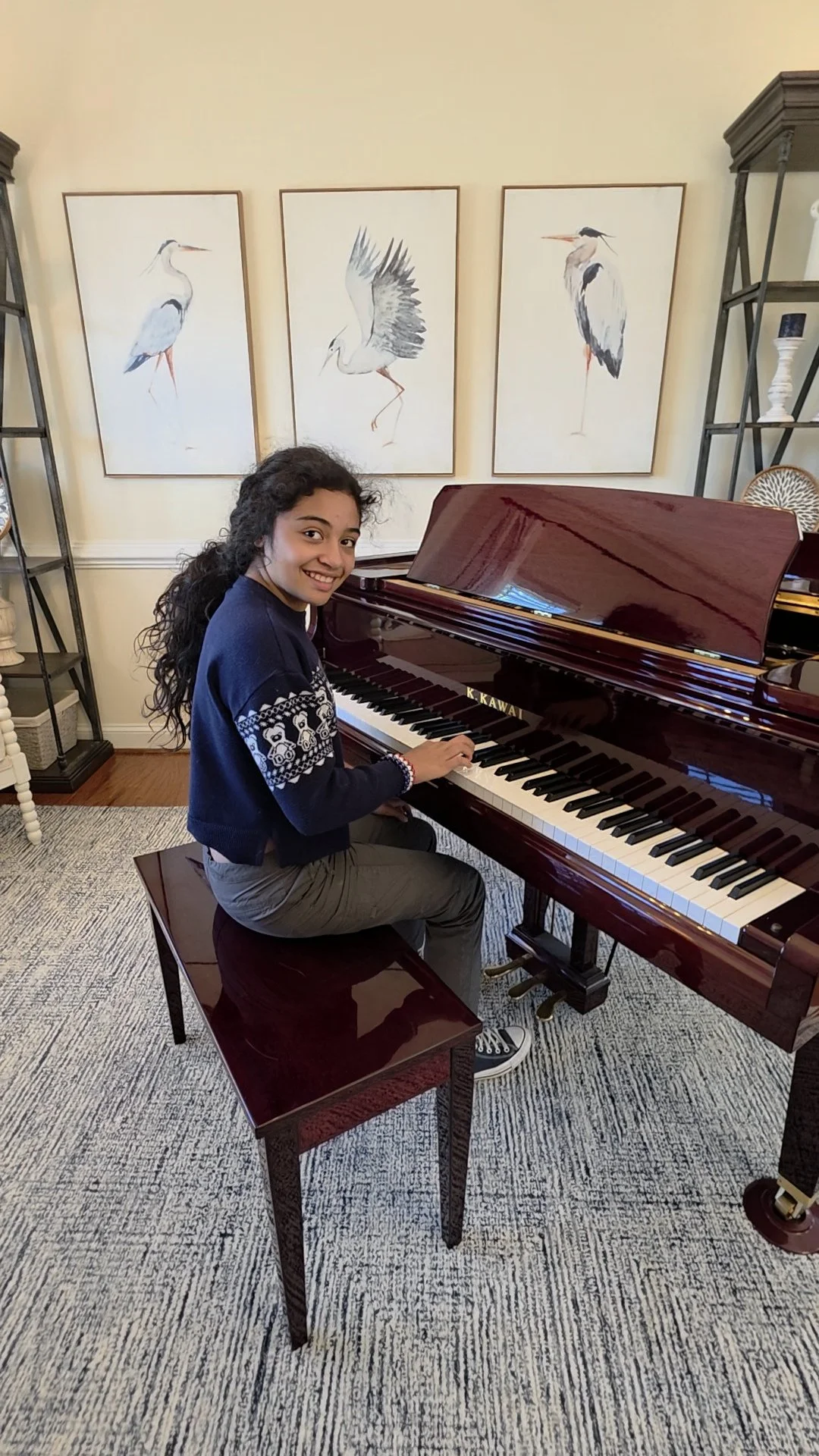 A young girl with dark curly hair sitting on a wooden piano bench, playing a polished grand piano with a smile. Behind her, three framed watercolor paintings of herons hang on a cream-colored wall. The room features wooden flooring and a patterned rug.