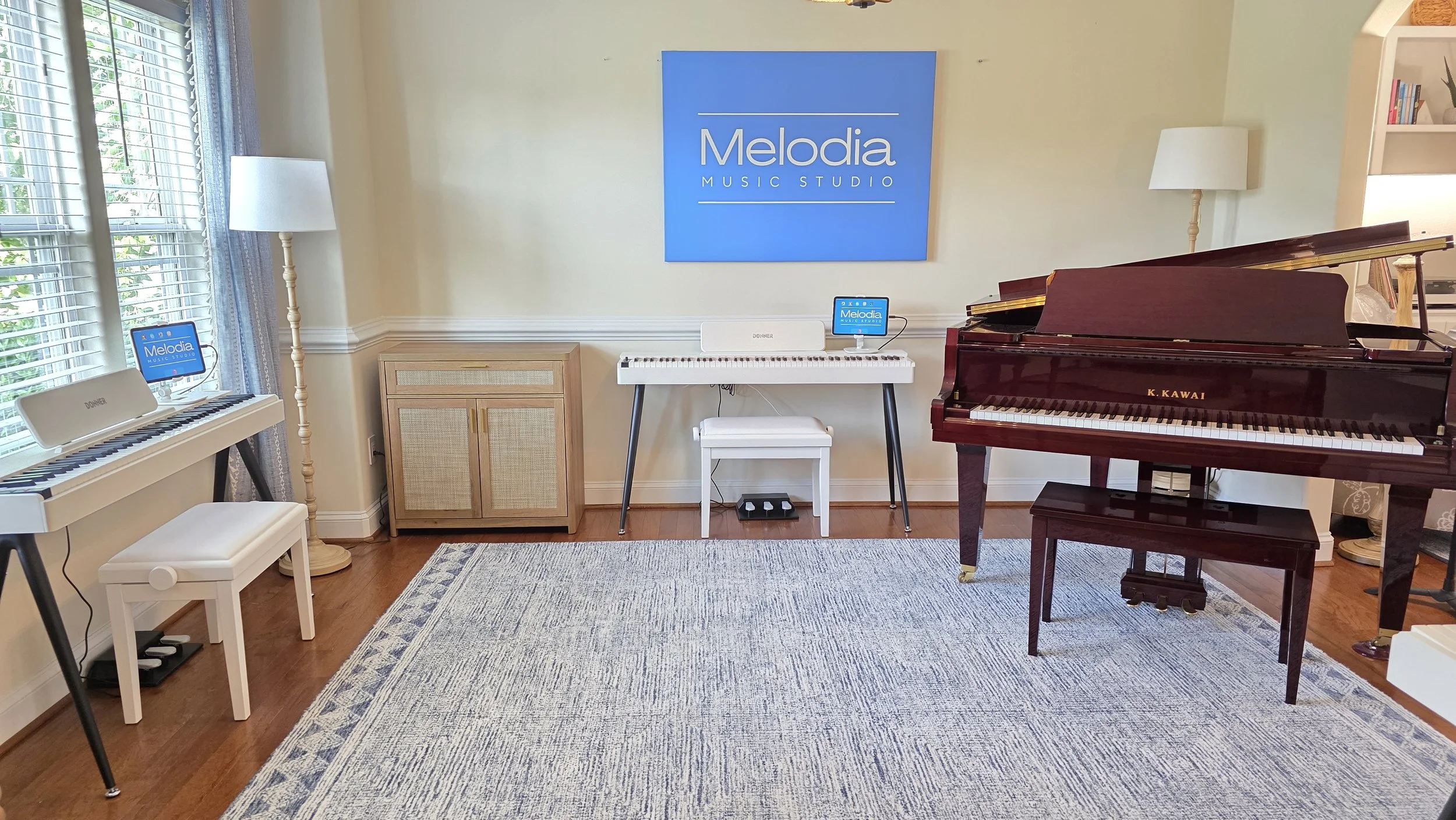 A music studio room featuring a red Kawai grand piano, an electronic keyboard, a digital tablet with Melodia Music Studio branding, a small speaker, a tall floor lamp, a wooden cabinet, a bookshelf, and a rug on hardwood floors with a window on the left side.