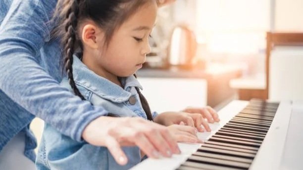 A young girl with braids learning to play piano with guidance from an adult.