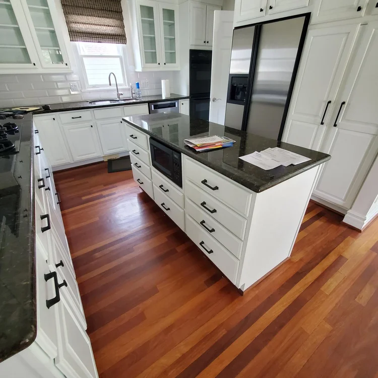 Clean, modern kitchen with white cabinets, black hardware, a black kitchen island with a microwave, hardwood floors, a built-in refrigerator, and a window above the sink.