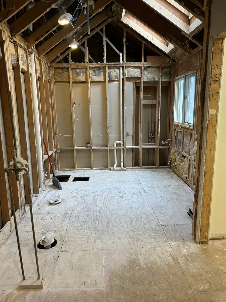 Room under renovation with exposed wooden framing, pipes, and a window, with construction tools on the floor.