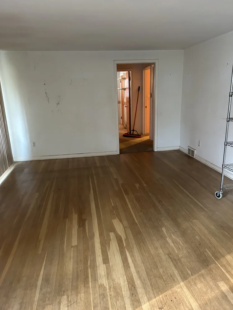 Empty room with hardwood floors, white walls, and a sliding glass door, with cleaning tools visible through a doorway in the background.