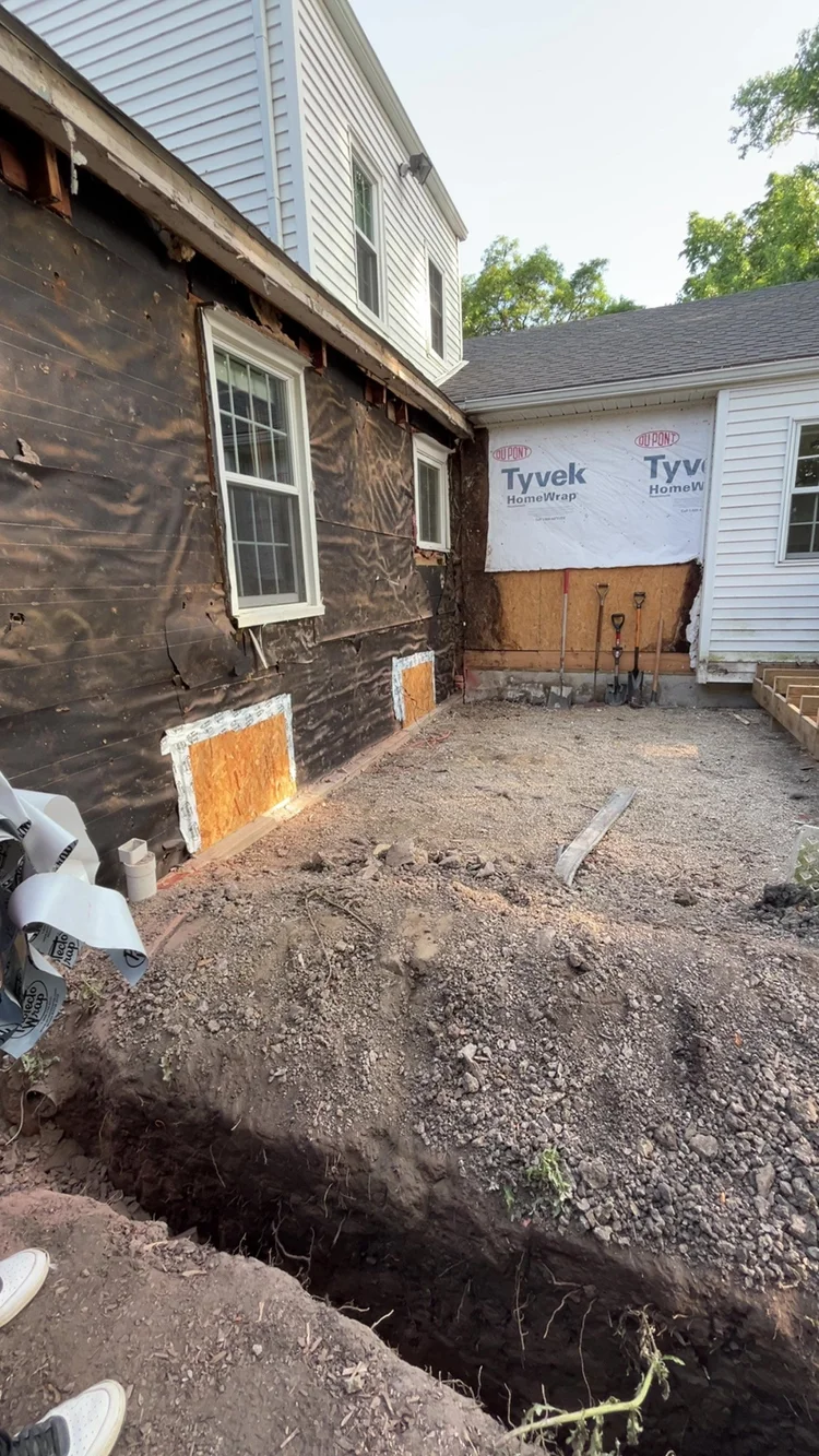 A house undergoing exterior renovation with part of the wall stripped down, showing black weather-resistant barrier and insulation, with a small trench dug in the yard for utility work.