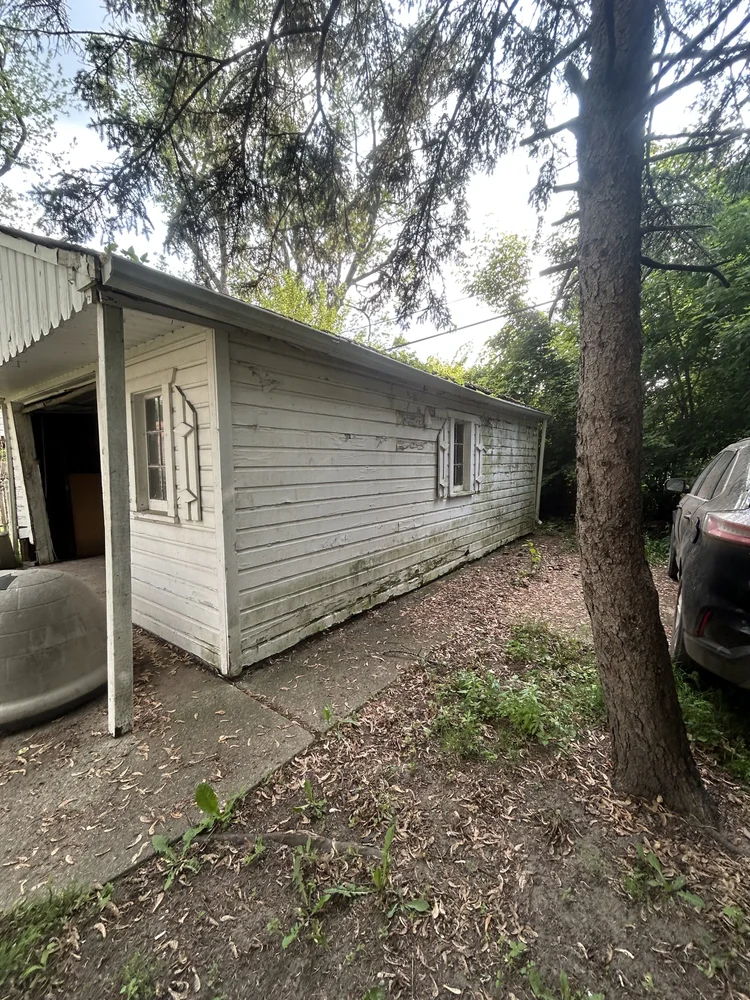 An old, weathered wooden house with peeling white paint, two small windows with security bars, and a concrete pathway surrounded by dirt, grass, and trees.