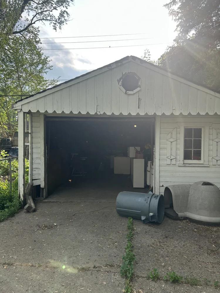 A small, white wooden garage with an open door, various items inside, a round vent above the garage door, and a plastic trash can and a doghouse outside on a dirt driveway with some weeds.