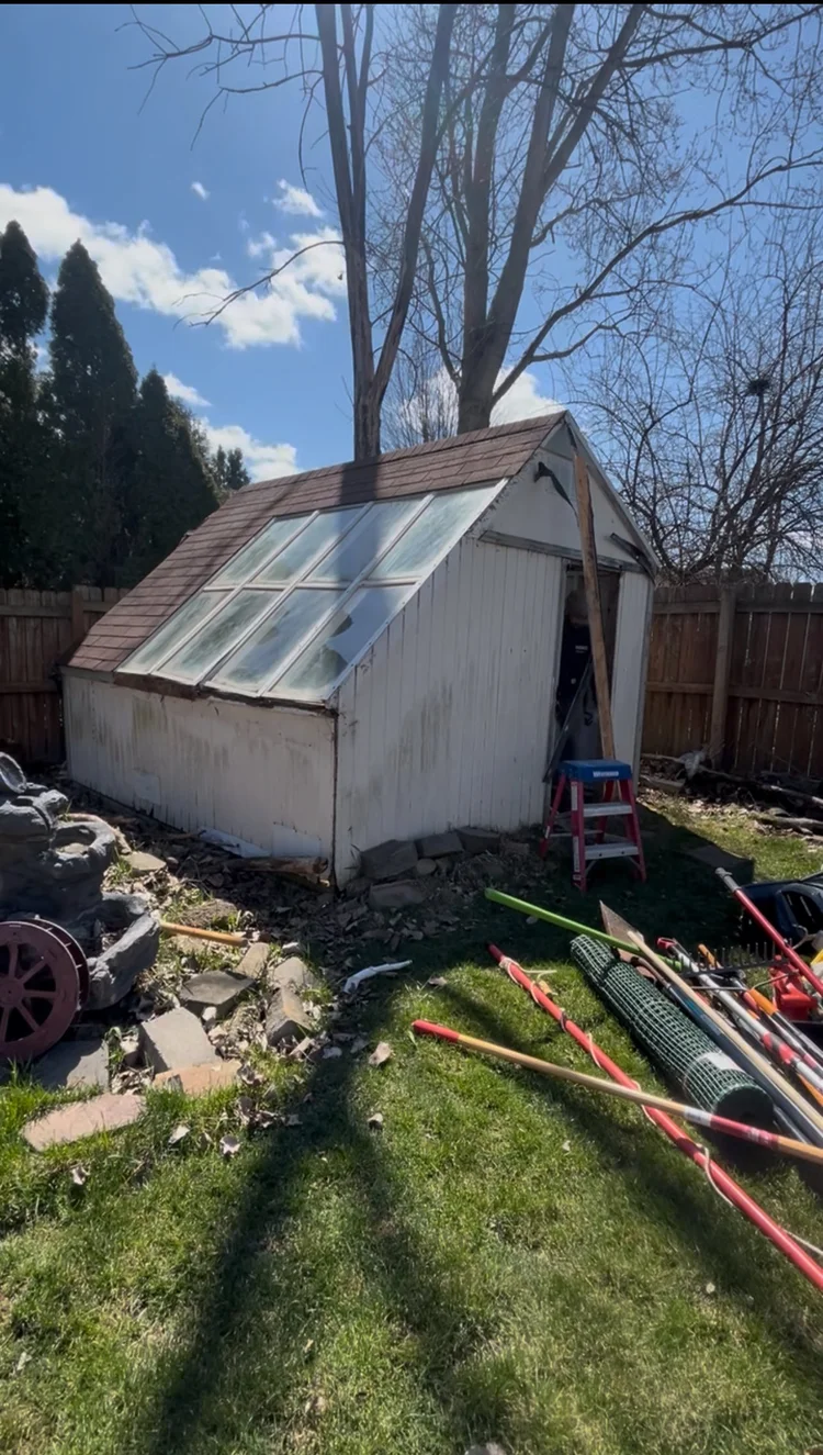 A small white wooden garden shed with a sloped glass roof, surrounded by tools and gardening equipment, in a backyard with a fence, trees, and a partly cloudy sky.