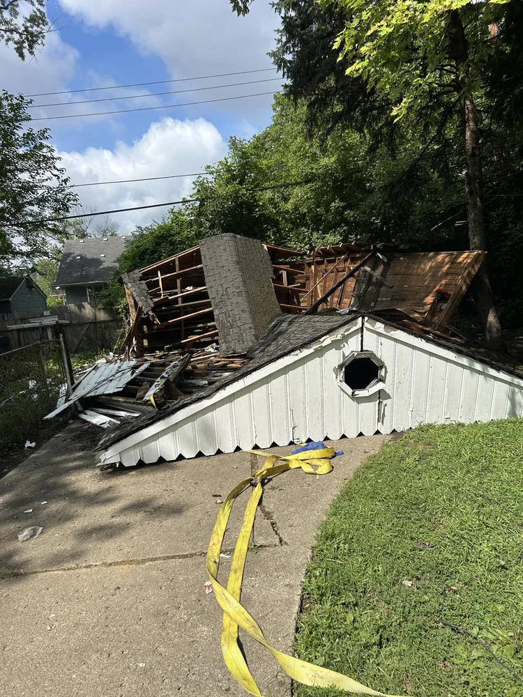 A damaged house with part of the roof collapsed, showing wooden beams and interior structure, lying on the ground. A yellow strap is on the concrete driveway in front of the house, with green grass and trees surrounding the area.