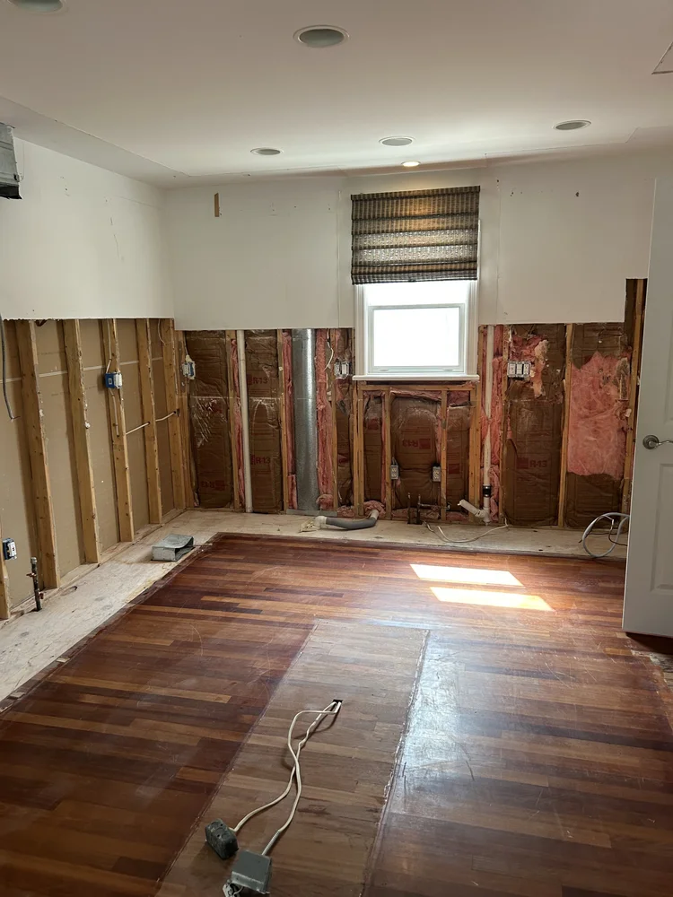 Room under renovation with exposed wall studs, insulation, electrical wiring, and partially completed hardwood floor, with a window covered by a striped window shade.