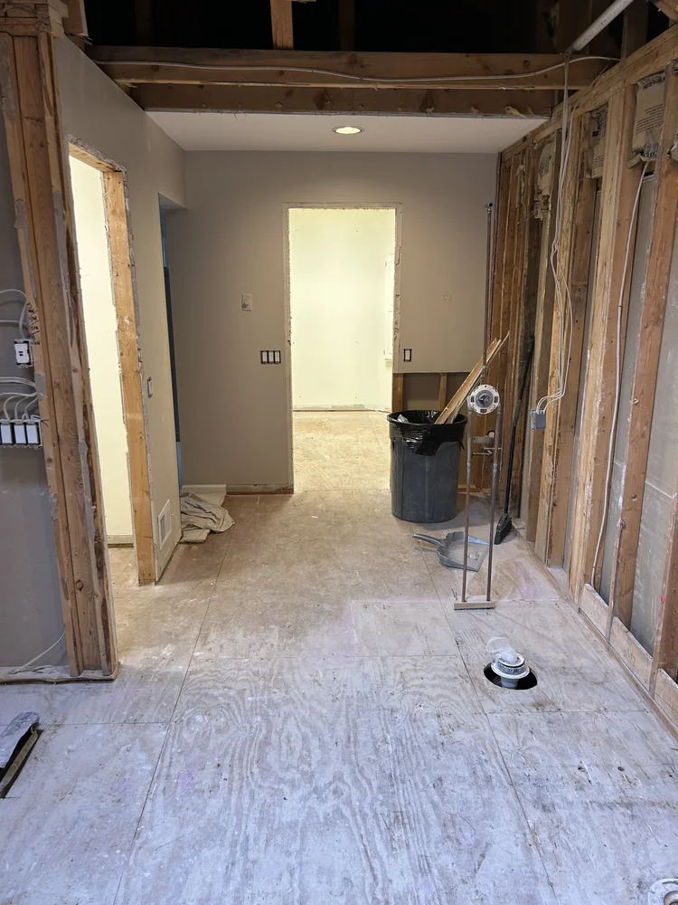 Interior view of a room under construction or renovation, with exposed wooden wall framing, unfinished drywall, construction tools, and a trash can.