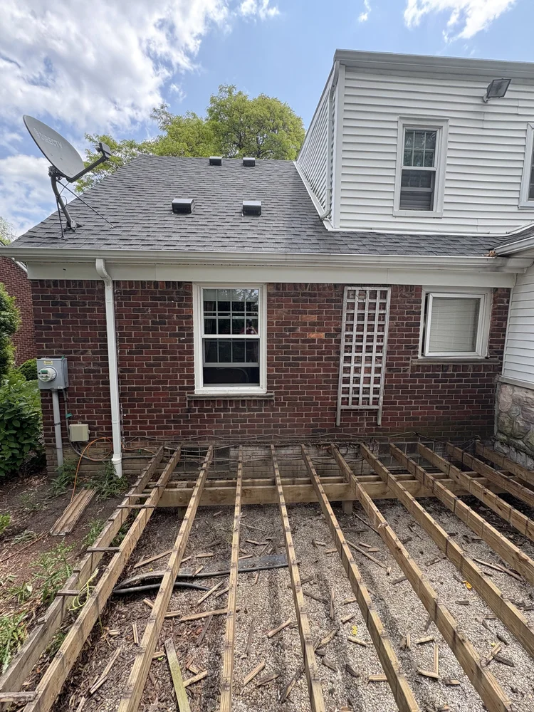 Backyard with a partially constructed wooden deck, a brick house with white siding on upper levels, two windows, a satellite dish, and a cloudy sky.