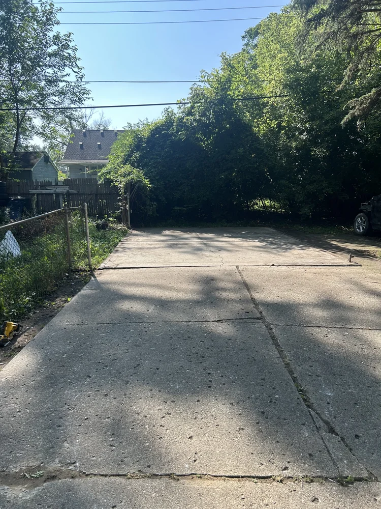 A paved driveway with a small gate on the left, trees and a house in the background, and a parked car on the right.