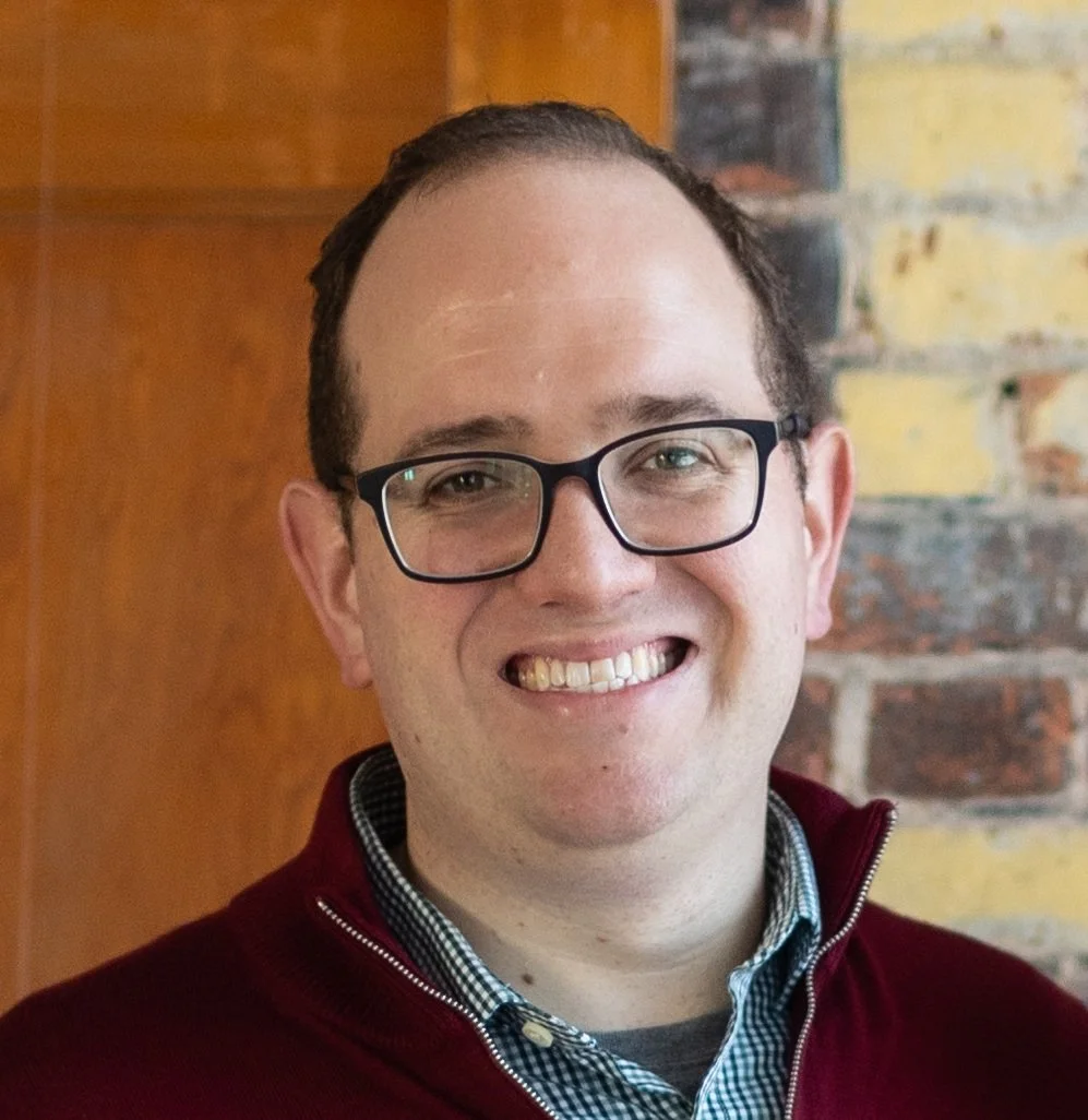 A man with glasses smiling, wearing a maroon sweater and checkered shirt, standing in front of a brick wall and wooden panel.