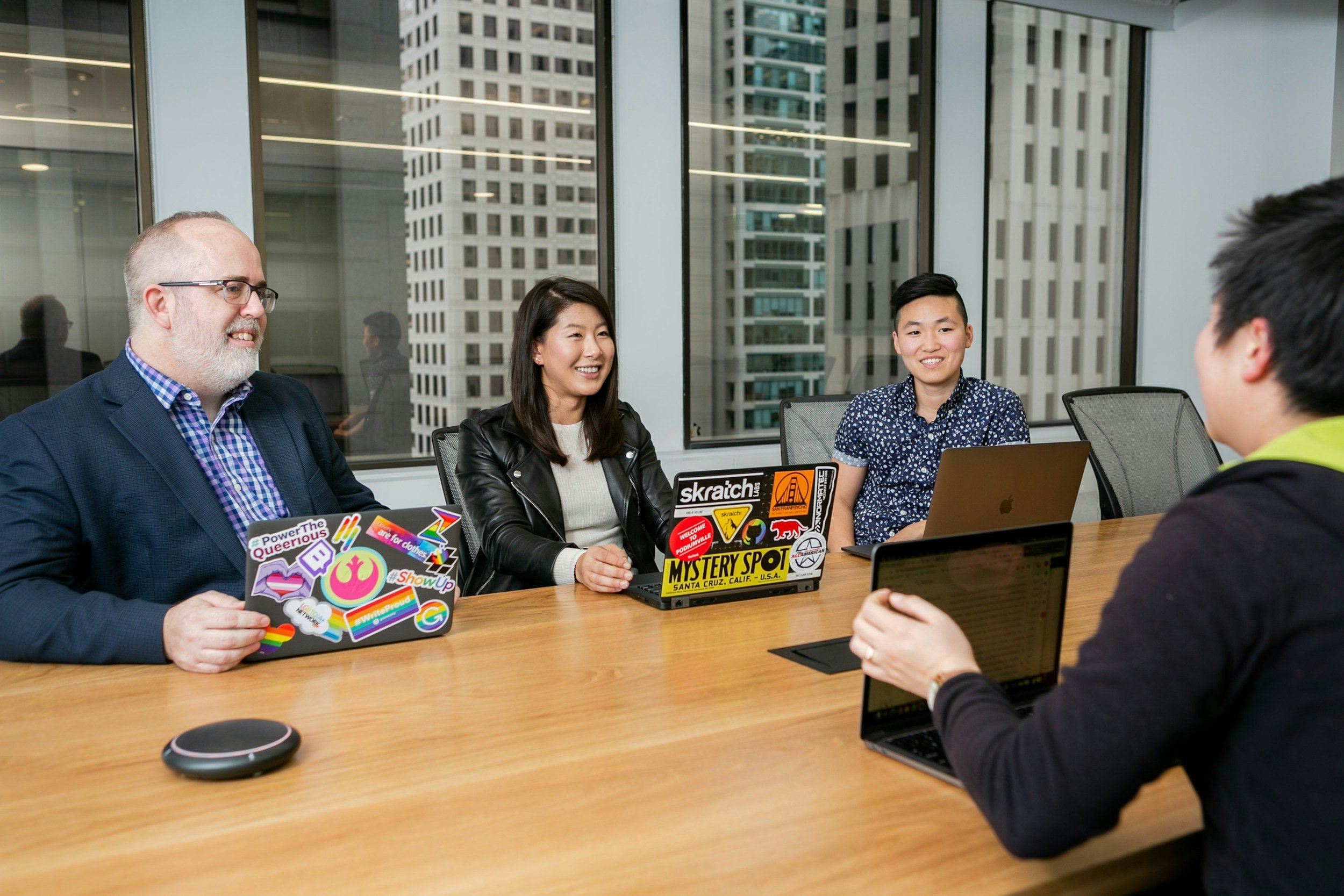 Four coworkers sitting at a conference table in a modern office, engaged in a discussion. They have laptops decorated with stickers, and there are large windows with city buildings in the background.