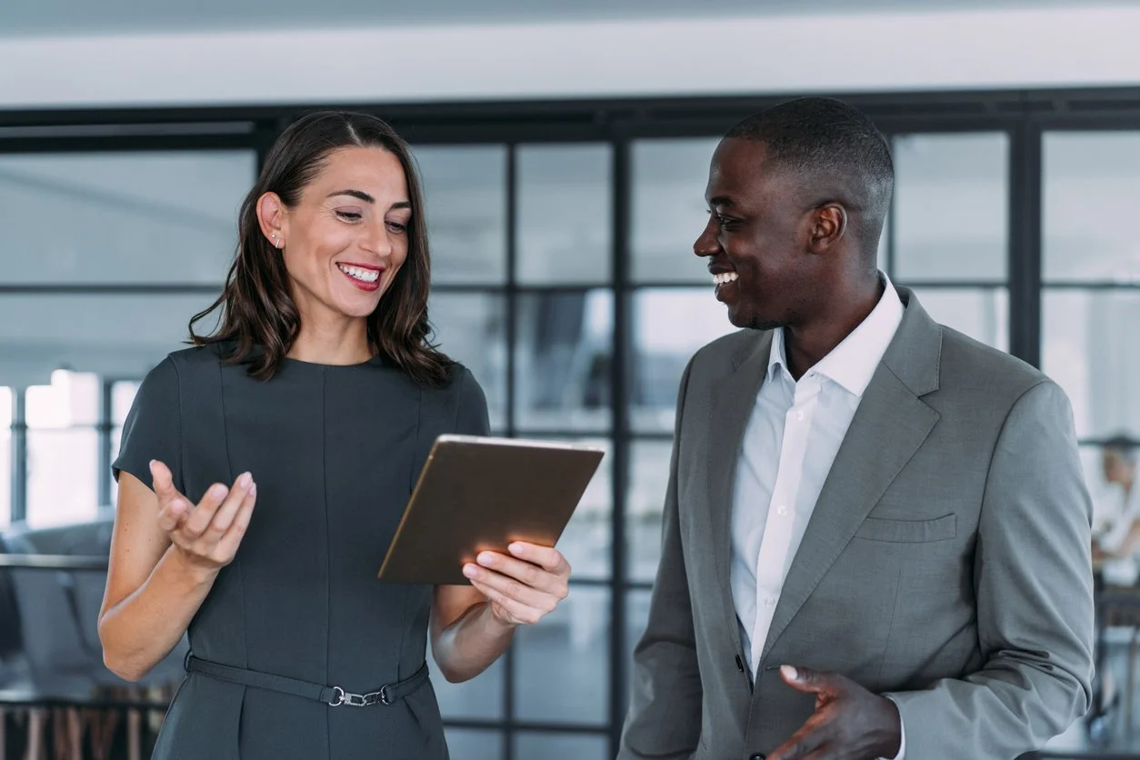 Two professionals, a woman and a man, smiling and looking at a tablet in an office setting.