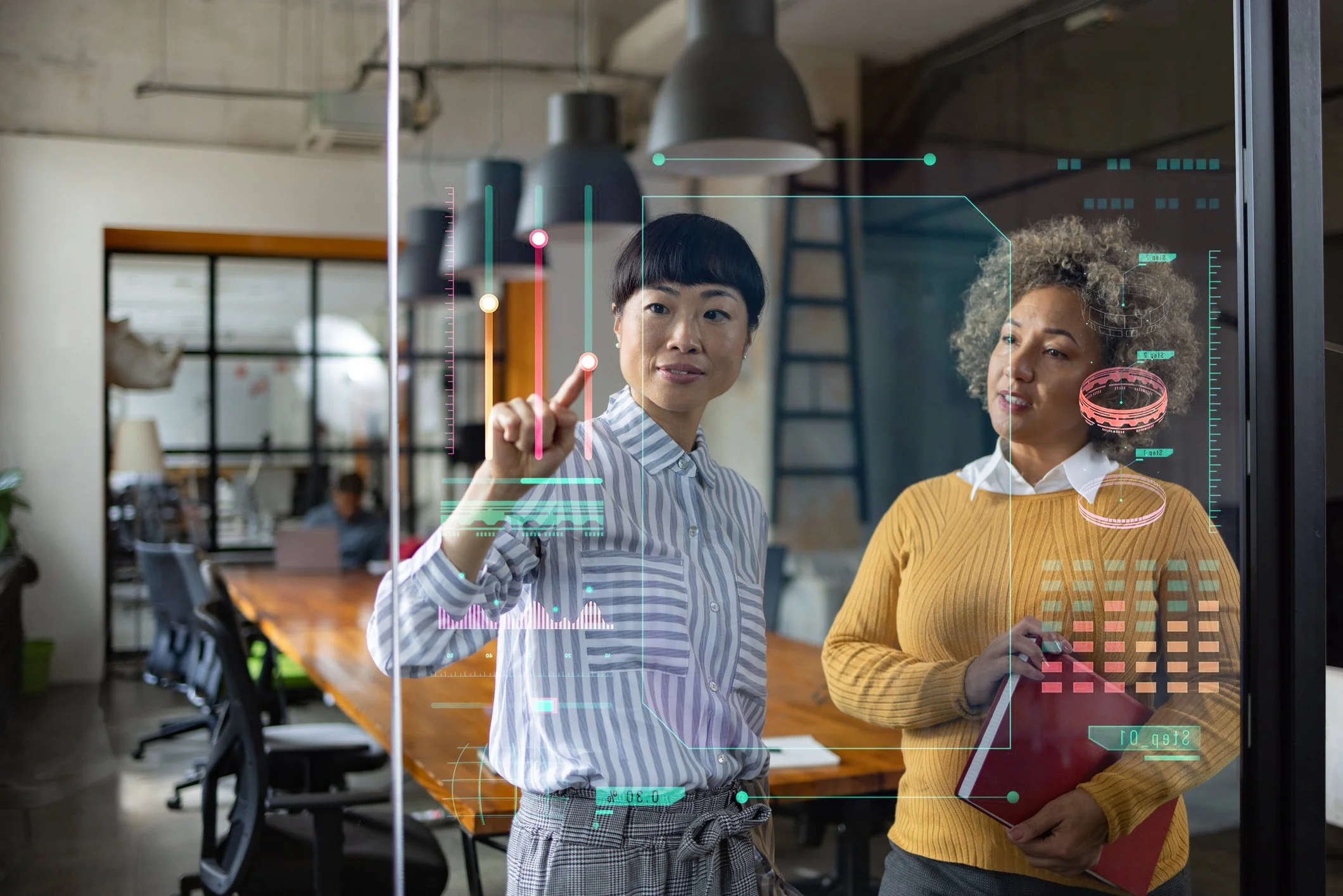 Two women in an office look at a transparent digital interface with charts and graphs. One woman points at the interface while the other holds a notebook.