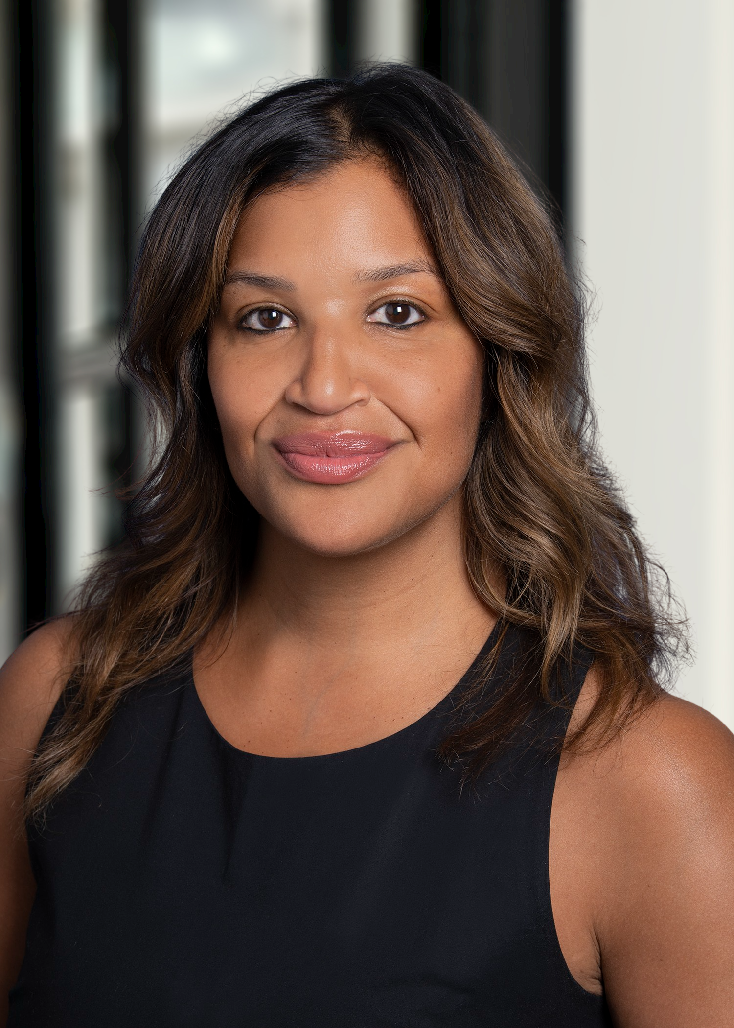 A woman with shoulder-length wavy hair, wearing a sleeveless black top, smiling softly, standing indoors near large windows.