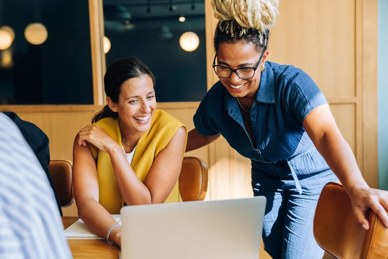Two women sharing a laugh while looking at a laptop in a casual meeting.