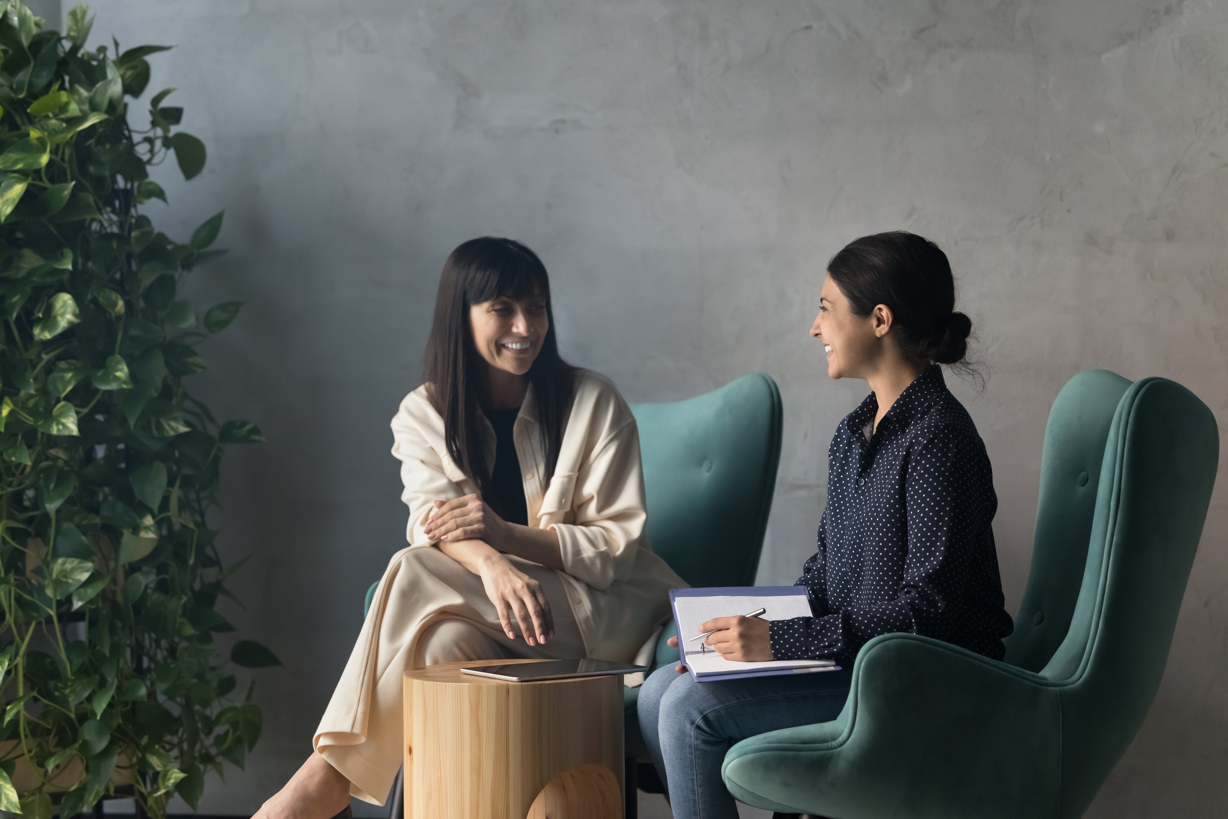 Two women sitting and talking in a modern office, one holding a clipboard with a pen, both smiling.