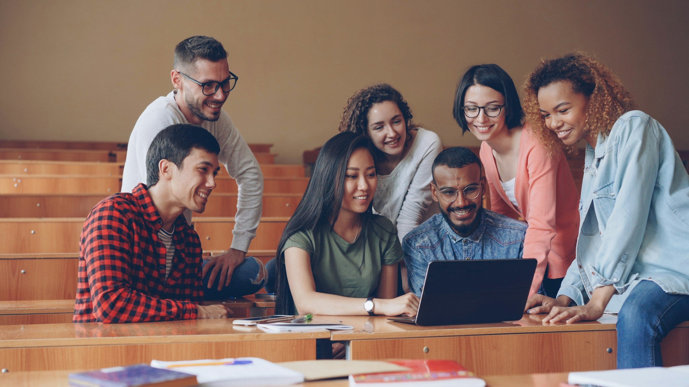 Group of diverse young students gathered around a laptop in a classroom, smiling and looking at the screen.