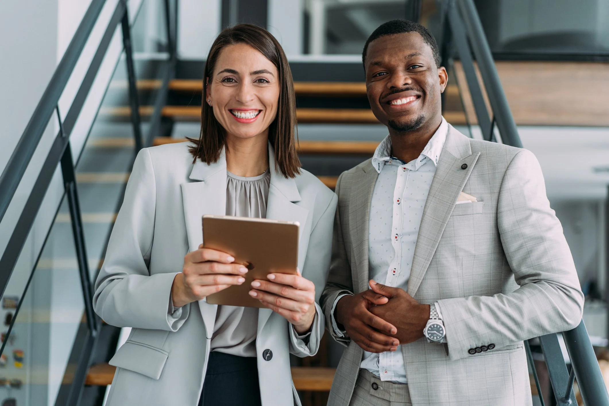 Two professionally dressed people, a woman and a man, smiling and standing close together indoors, with a staircase in the background. The woman is holding a tablet.