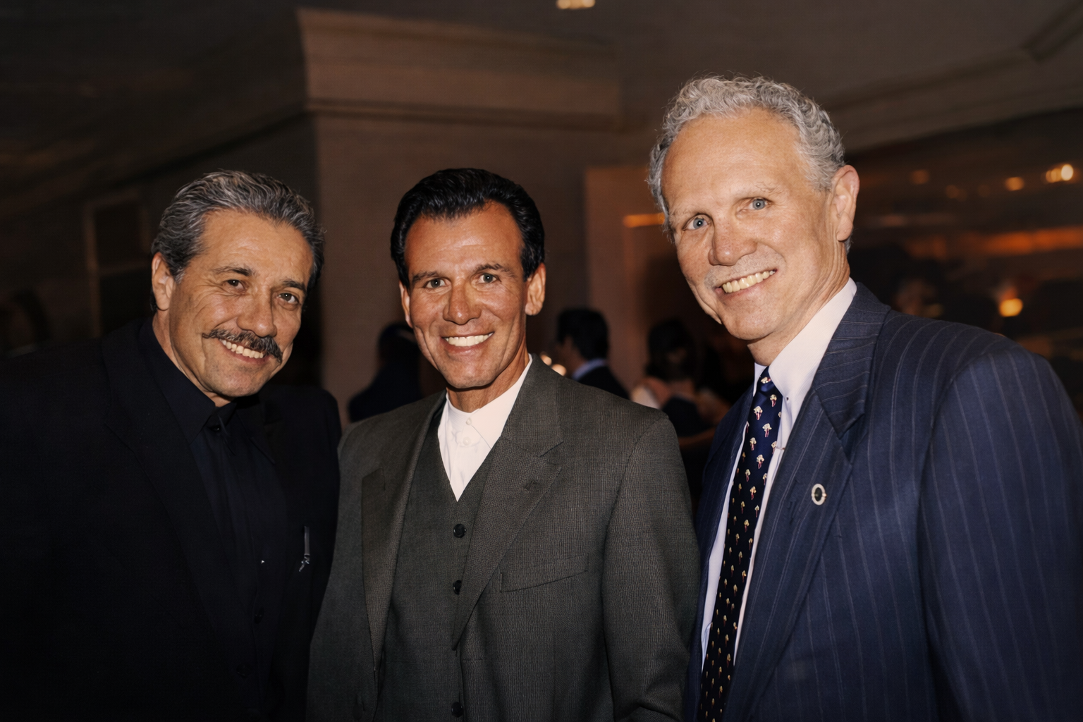 Three men in formal attire smiling at the camera at an indoor event with warm lighting and blurred background of other attendees.
