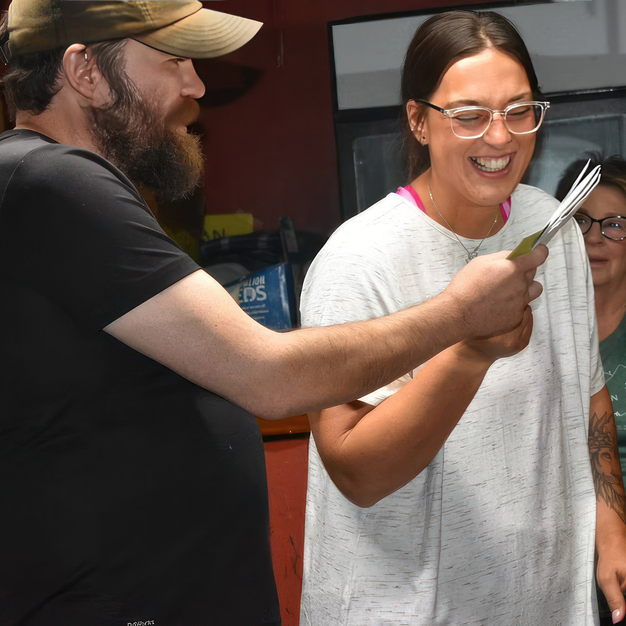 A bearded man in a baseball cap presenting a card to a smiling woman with glasses. Another woman in glasses looks on in the background.