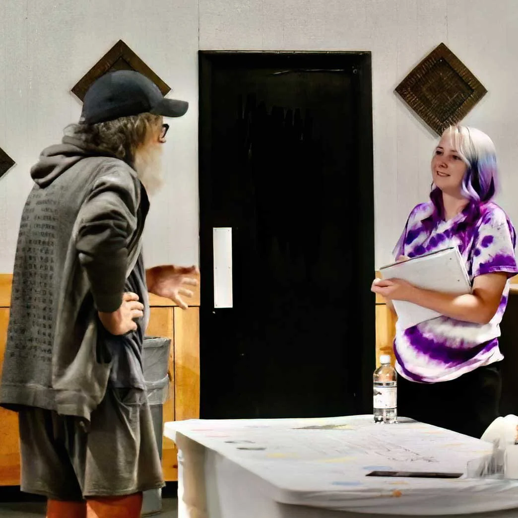 A woman with purple and white hair holding a notebook or clipboard talking to a man with a beard, glasses, hat, and casual clothing. They are indoors near a table with a water bottle, with a dark door behind them and decorative wall accents.