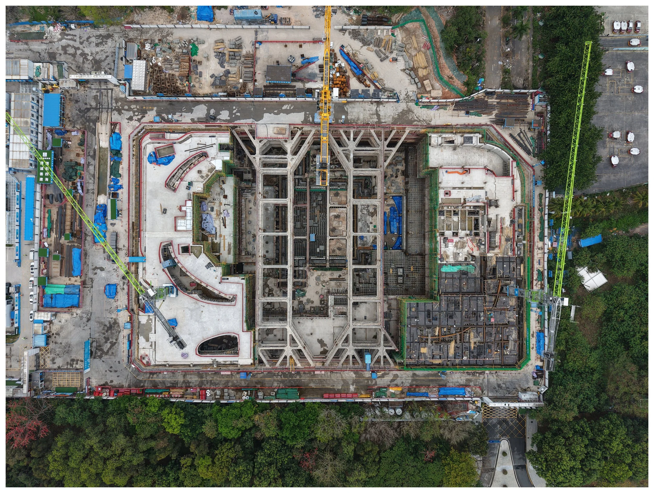 Aerial view of a construction site with cranes, concrete structures, and various building materials, surrounded by trees and parking lot.
