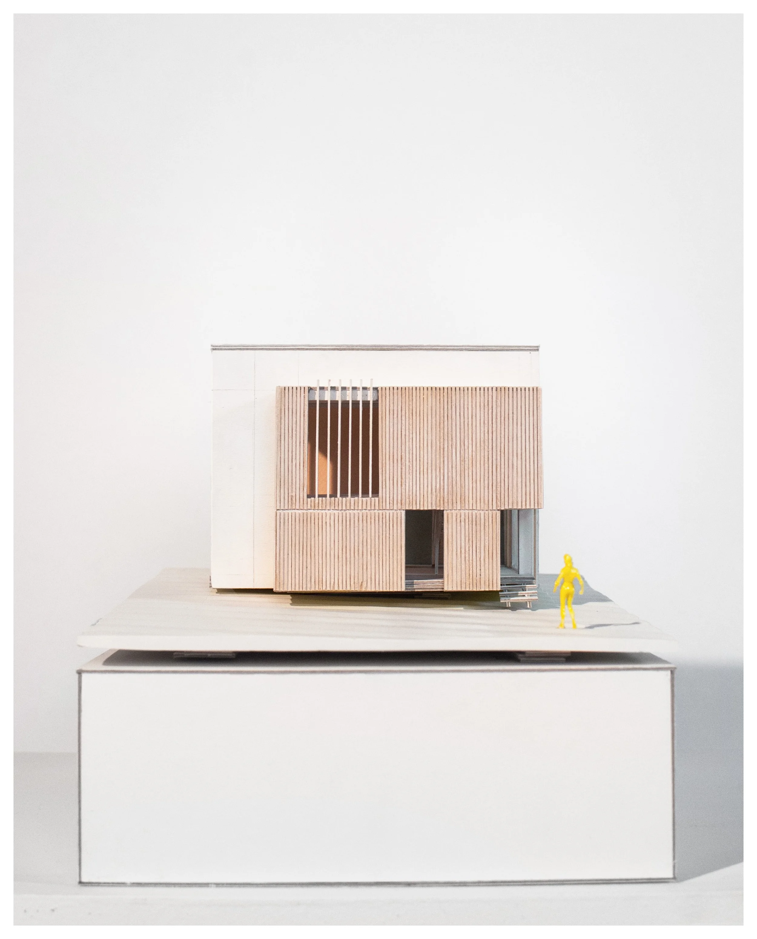 Architectural model of a modern house with wooden exterior, placed on a white display stand, with a small yellow human figure for scale.