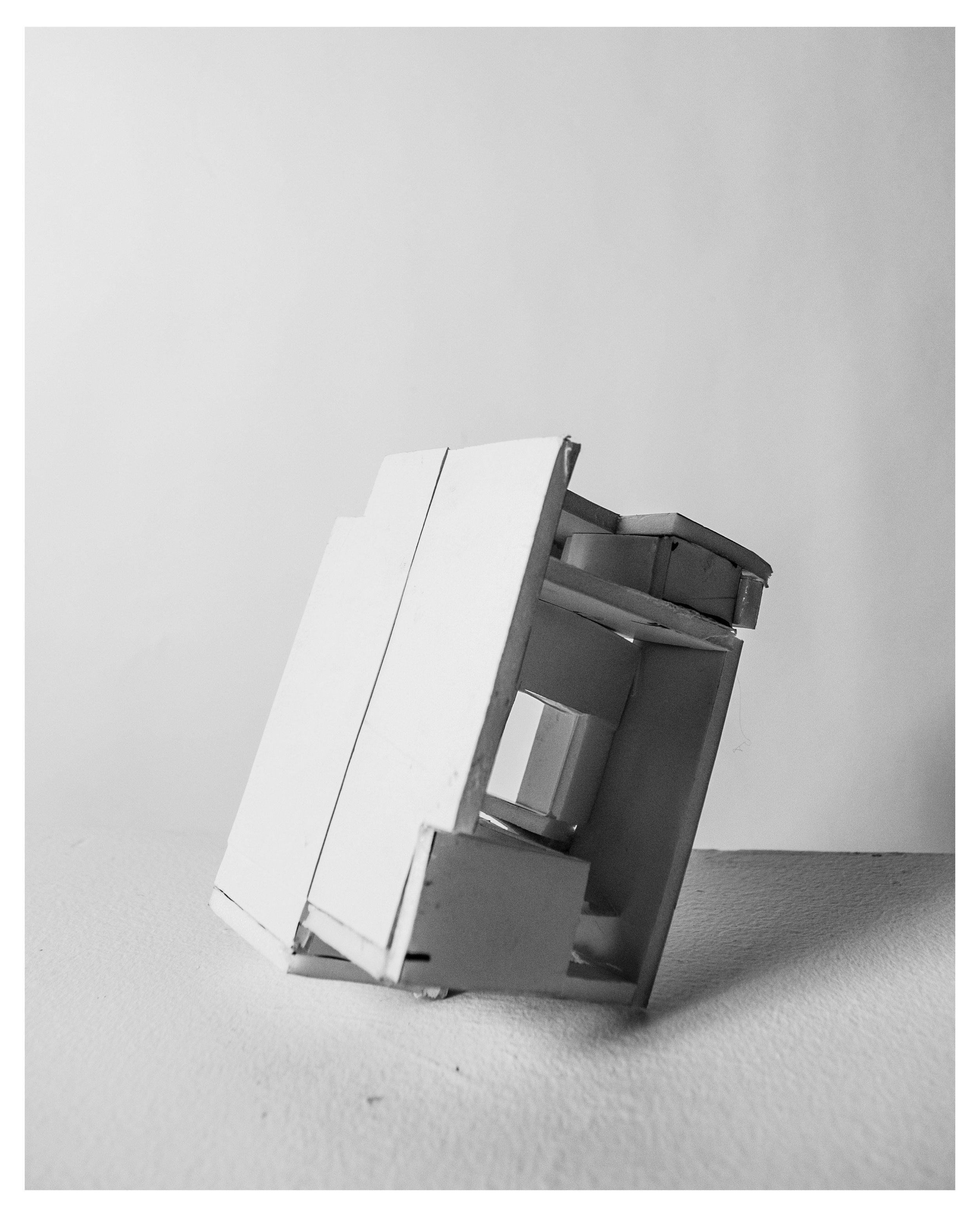 A small, tilted white and gray model of a house made of cardboard placed on a flat surface against a plain wall.