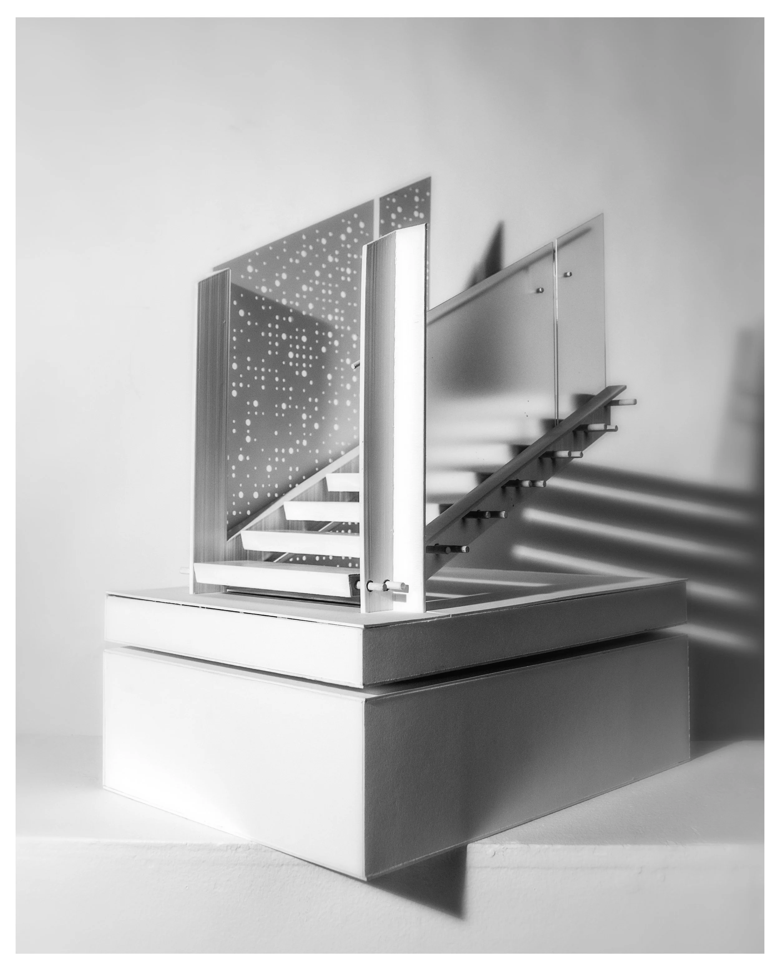 A modern, minimalist staircase with perforated metal risers, enclosed by clear glass panels, mounted on a rectangular platform with a shadow cast on the wall behind.
