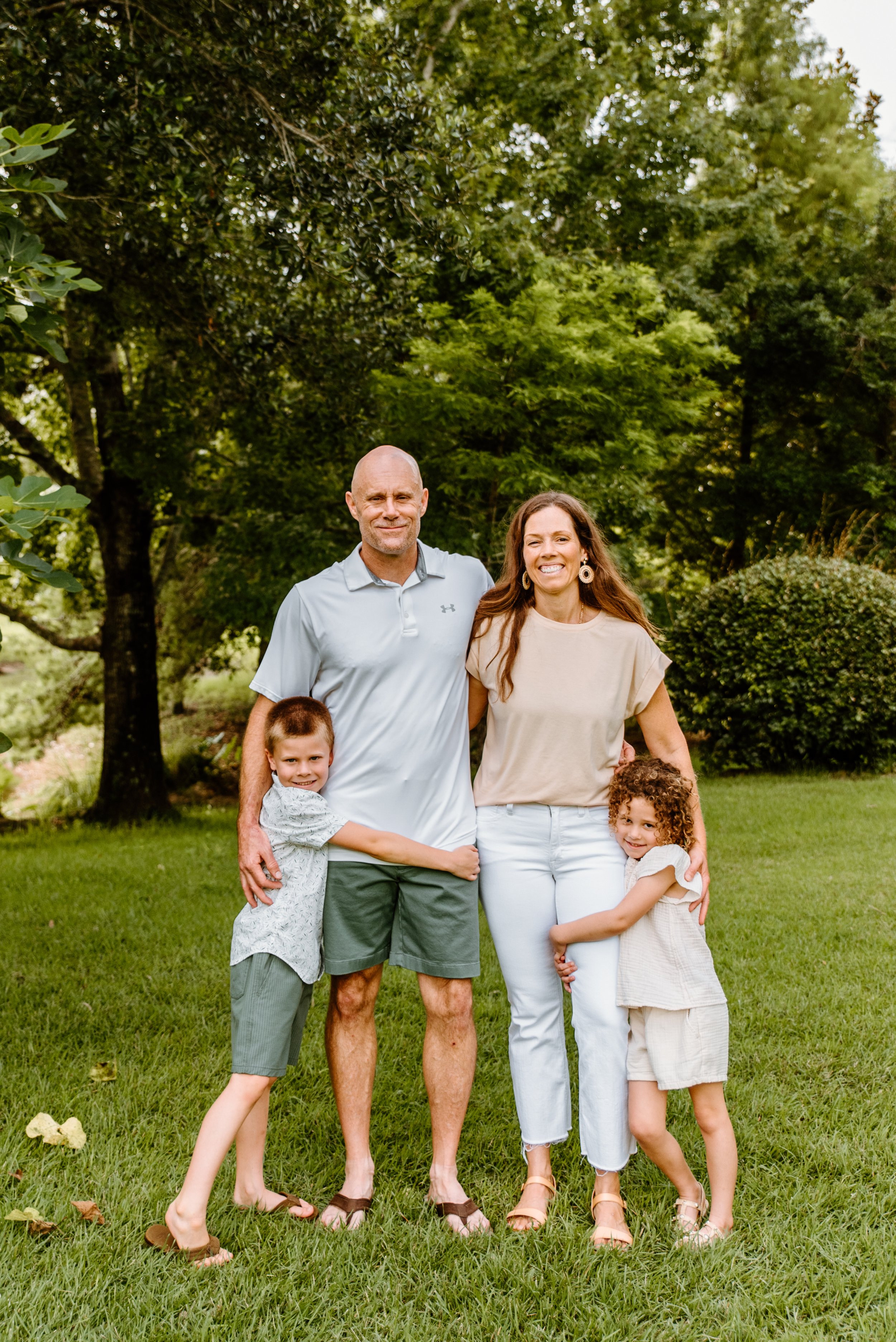 A family of four standing on a grassy lawn with green trees in the background, smiling at the camera. The father is bald, wearing a light gray polo shirt and green shorts. The mother has long brown hair, wearing a beige top and white pants. The son has short brown hair, wearing a patterned shirt and green shorts. The daughter has curly hair, wearing a light-colored dress.